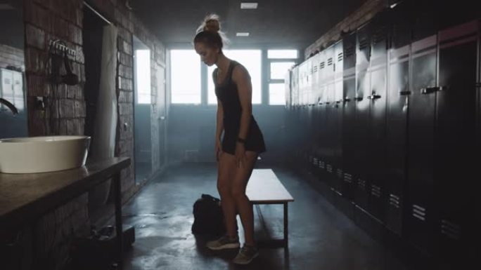 摄像机跟随欧洲运动女子进入黑暗的健身房更衣室，坐着感到疲倦和沮丧的慢动作。
