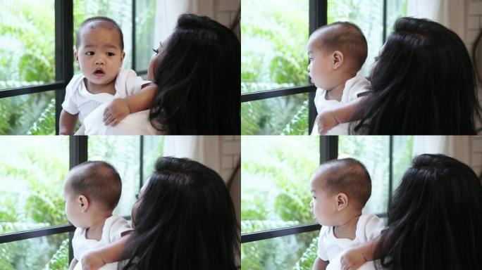 4k慢动作镜头，亚洲男婴凝视着窗户玻璃，而他的母亲抱着现代阁楼房子，家庭和生活方式的概念