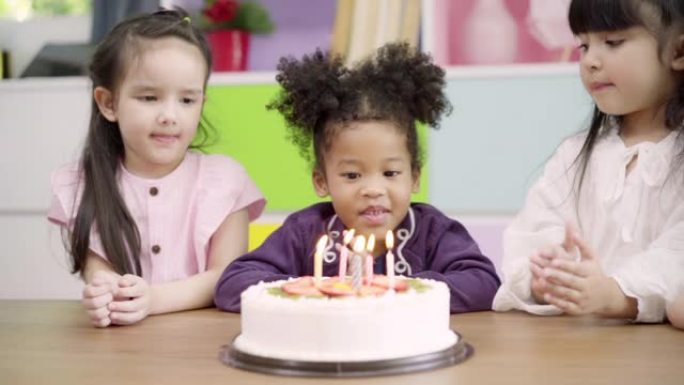 慢动作-孩子们在教室里庆祝生日聚会，多民族的年轻女孩快乐地许愿在学校的生日蛋糕上吹灭蜡烛。孩子们在学