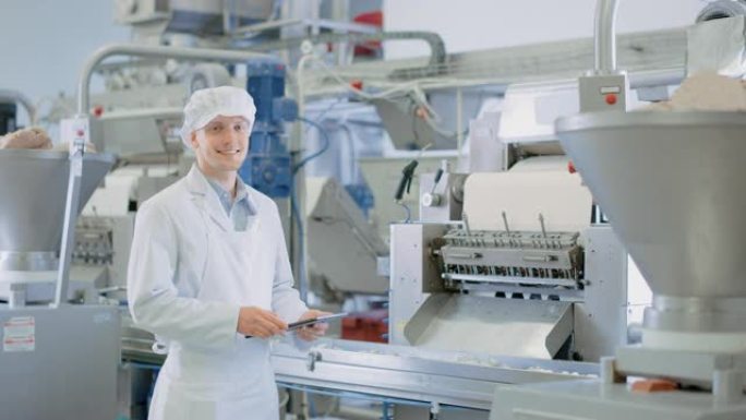 年轻的男性质量主管或食品技术员正在饺子食品工厂检查自动化生产。员工使用平板电脑工作。他看着镜头微笑。