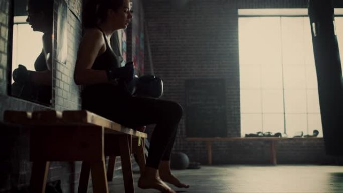 美丽健康的黑发跆拳道运动员坐在阁楼健身房的长凳上，用激励海报。她屏住呼吸，脱下战斗手套。经过紧张的训