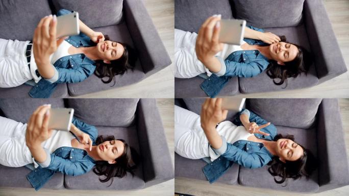 迷人的女孩躺在家里的沙发上用智能手机相机自拍