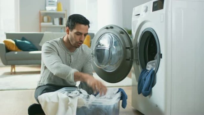 穿着灰色牛仔裤和套头衫的英俊微笑的年轻人坐在家里的洗衣机前。他给洗衣机装上脏衣服。明亮宽敞的客厅，室