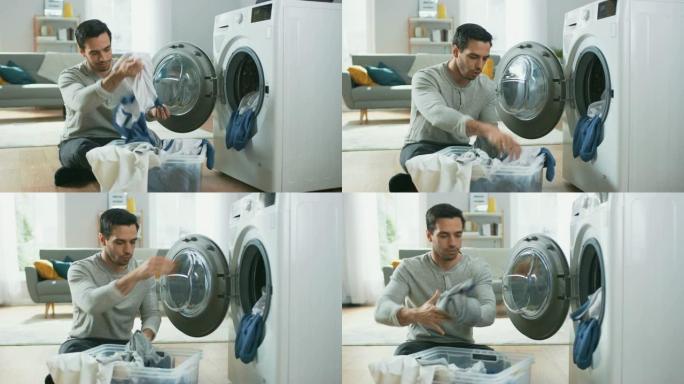 穿着灰色牛仔裤和套头衫的英俊微笑的年轻人坐在家里的洗衣机前。他给洗衣机装上脏衣服。明亮宽敞的客厅，室