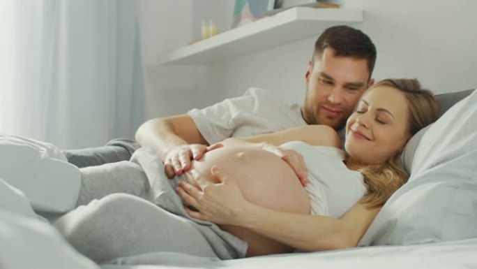 幸福的年轻夫妇在床上拥抱在一起，年轻的女人怀孕了，充满爱心的丈夫温柔地抚摸着她的肚子。相机聚焦在腹部