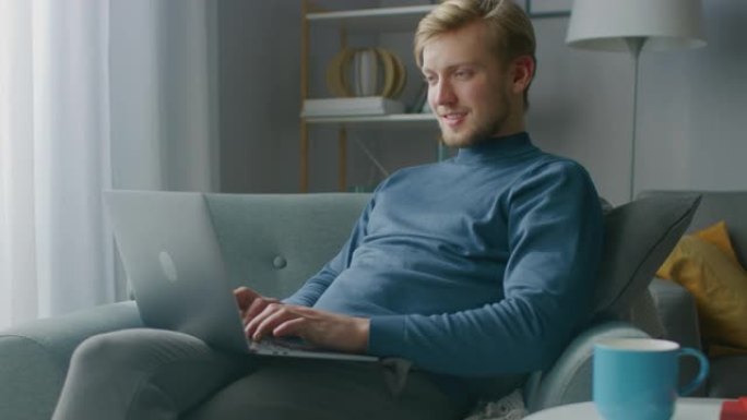 英俊的金发小伙子在笔记本电脑上工作的肖像，而坐在他舒适的客厅的椅子上。创意自由职业者在家放松，浏览互