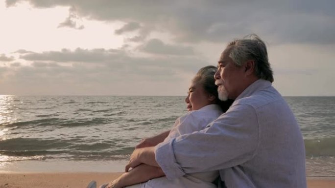 老年夫妇一起坐在沙滩上，看着海洋上的日落。老年夫妇在晴天，高级假期，假期在海边放松