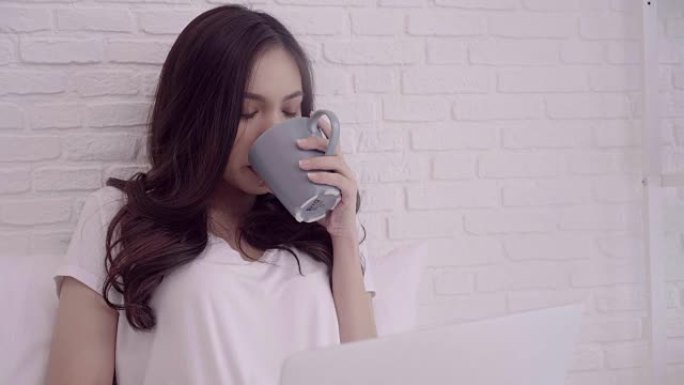 美丽迷人的亚洲妇女的肖像使用电脑或笔记本电脑拿着一杯温暖的咖啡或茶，而躺在床上放松时，在她的卧室在家