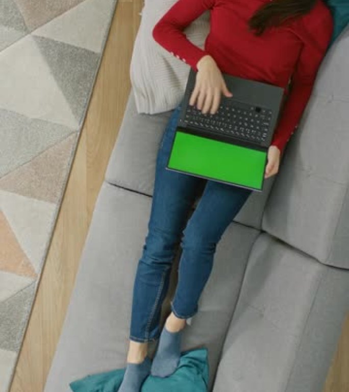 穿着红色套头衫的快乐年轻女孩躺在沙发上，使用带有绿色屏幕的笔记本电脑。舒适的客厅，室内现代。放大俯视
