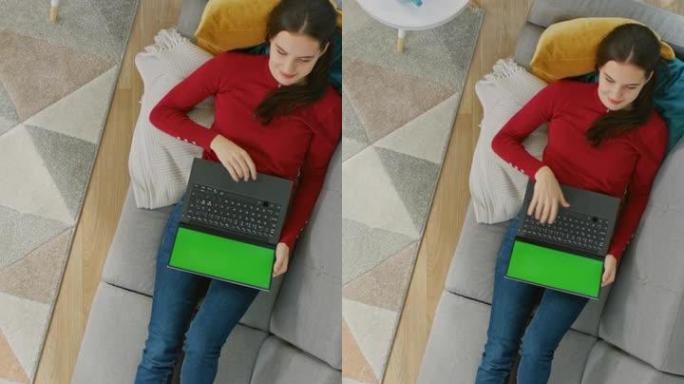穿着红色套头衫的快乐年轻女孩躺在沙发上，使用带有绿色屏幕的笔记本电脑。舒适的客厅，室内现代。放大俯视