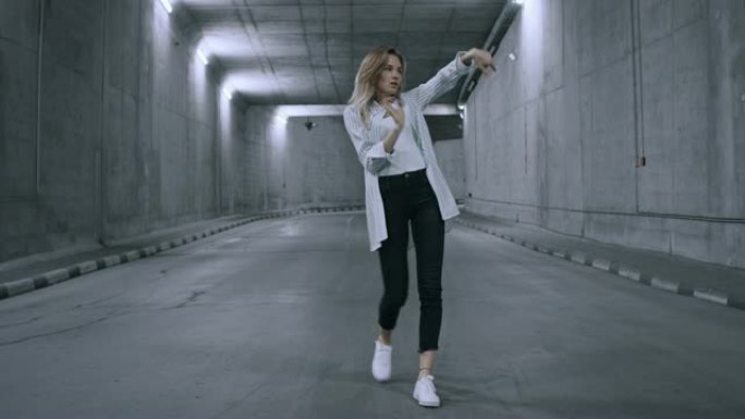 自信美丽酷的年轻金发女子正在点燃的混凝土隧道里积极跳舞。她穿着条纹白衬衫和深色牛仔裤。
