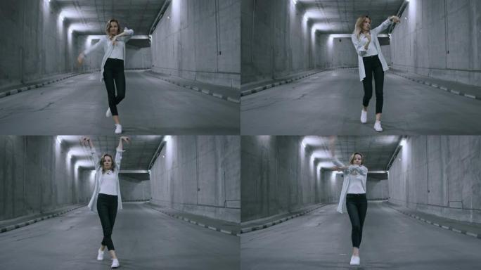 自信美丽酷的年轻金发女子正在点燃的混凝土隧道里积极跳舞。她穿着条纹白衬衫和深色牛仔裤。