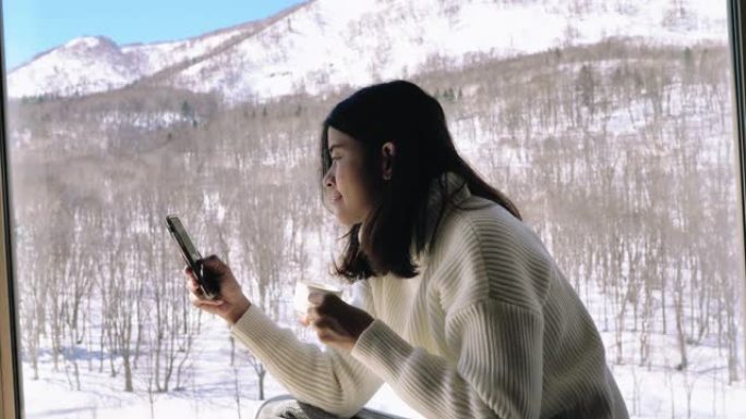 年轻的亚洲妇女坐在咖啡店里，在下雪时使用智能手机