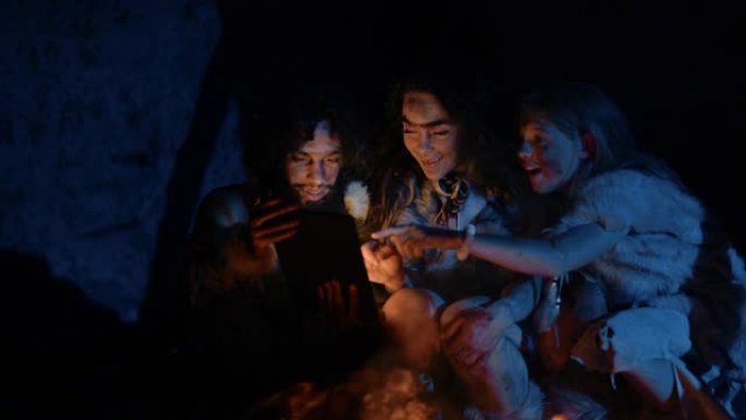 穿着兽皮的史前原始狩猎采集者部落在夜间在洞穴中使用数字平板电脑。尼安德特人或智人家庭浏览互联网，观看