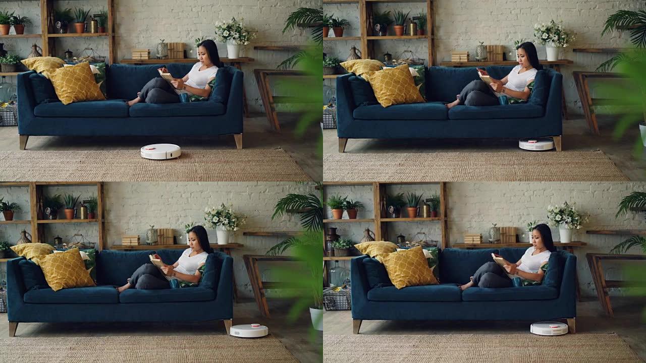 机器人真空吸尘器徘徊在现代阁楼风格的木地板上，在地毯上移动，而漂亮的亚洲女士正在看书，享受空闲时间。