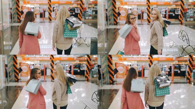 快乐的年轻女士的慢动作快乐的顾客在商场里一起散步，手里拿着五颜六色的袋子，然后转向相机微笑。购物狂和