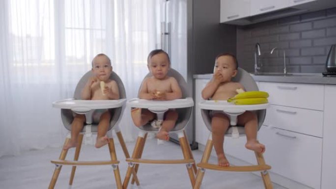 亚洲婴儿三胞胎坐在厨房的高脚椅上吃香蕉