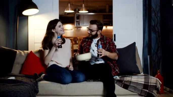快乐的年轻高加索浪漫情侣坐在一起在家放松的电影之夜，吃零食和饮料慢动作。