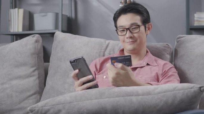 亚洲男子享受使用手机或智能手机进行在线购物，并持有信用卡在他的订单上付款。成人坐在家里客厅的沙发上进