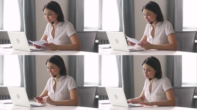 微笑的亚洲女商人戴着耳机说话看着笔记本电脑