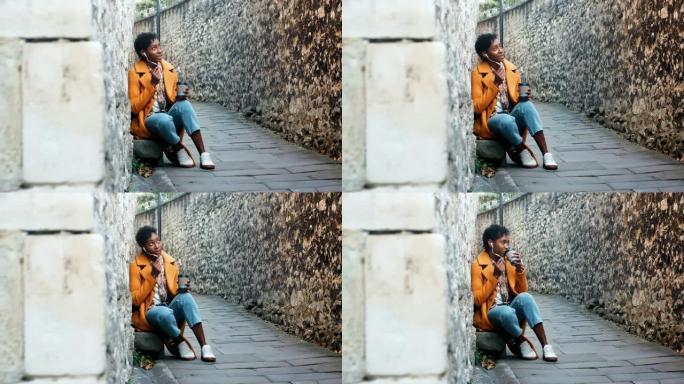 穿着黄色豌豆外套和蓝色牛仔裤的年轻女子坐在一个历史悠久的城市的小巷里，用耳机在智能手机上聊天，全长，