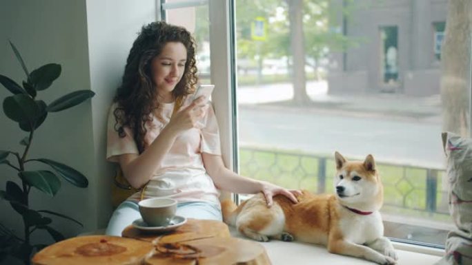 迷人的女人用智能手机坐在咖啡馆的窗台上，带着小狗