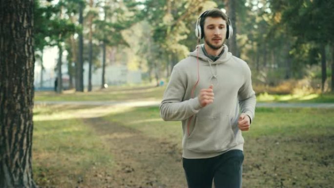 多莉拍摄了长着胡子的好男人，戴着耳机在公园里跑步，在阳光明媚的秋天听音乐。现代科技和体育理念。