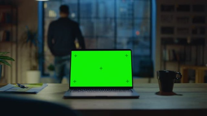 台式电脑，模拟绿屏站在现代创意办公室的桌子上。在后台，设计师使用智能手机看着窗外的夜城。放大镜头