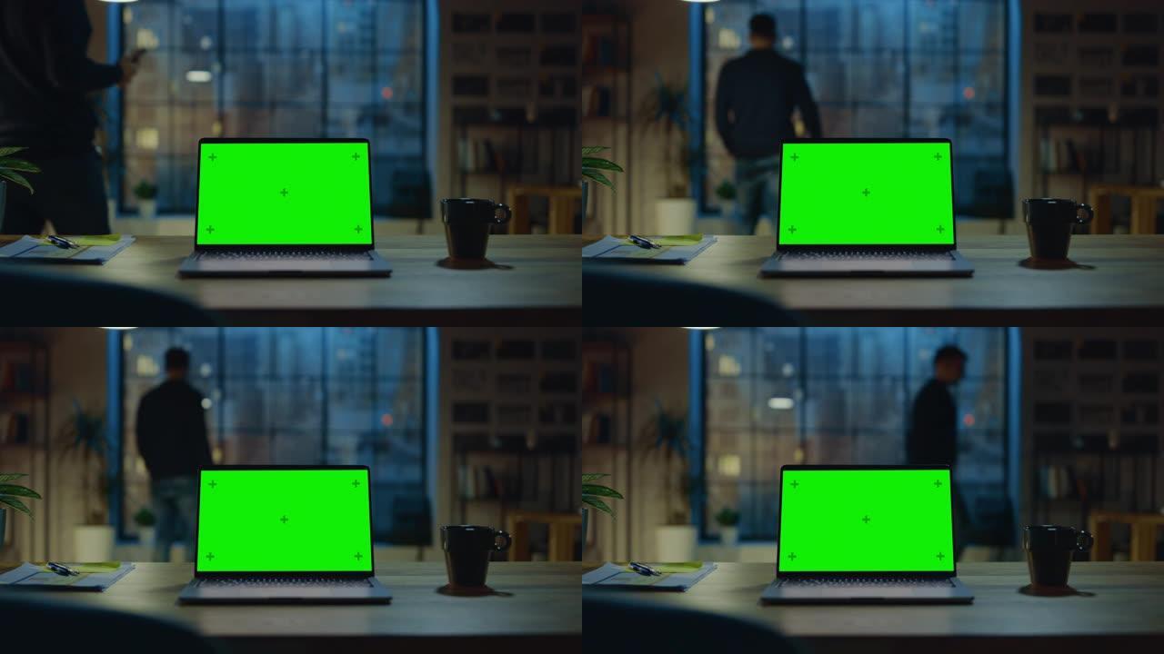 台式电脑，模拟绿屏站在现代创意办公室的桌子上。在后台，设计师使用智能手机看着窗外的夜城。放大镜头