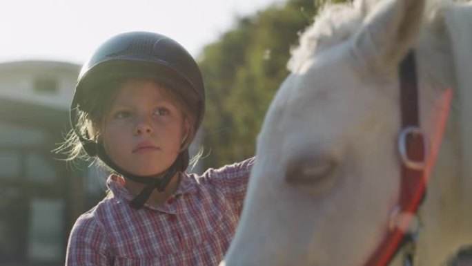 一个戴着骑师头盔的可爱的小女孩的真实特写镜头正在阳光下的马厩里爱抚着一匹白色小马马