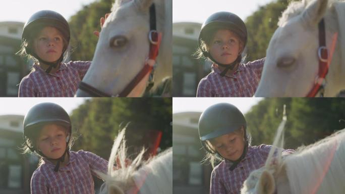 一个戴着骑师头盔的可爱的小女孩的真实特写镜头正在阳光下的马厩里爱抚着一匹白色小马马
