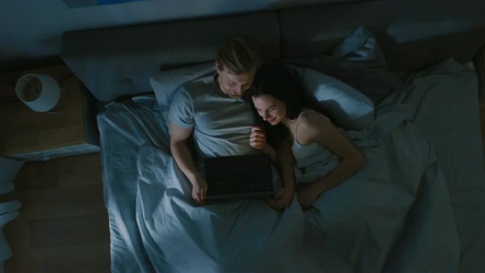 可爱的年轻夫妇晚上躺在床上，使用笔记本电脑，观看系列节目，阅读社交媒体，浏览互联网，交谈和娱乐。顶视
