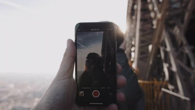 男性手中的智能手机正在拍摄艾菲尔铁塔慢动作顶部摆姿势的浪漫微笑女人的浪漫肖像。
