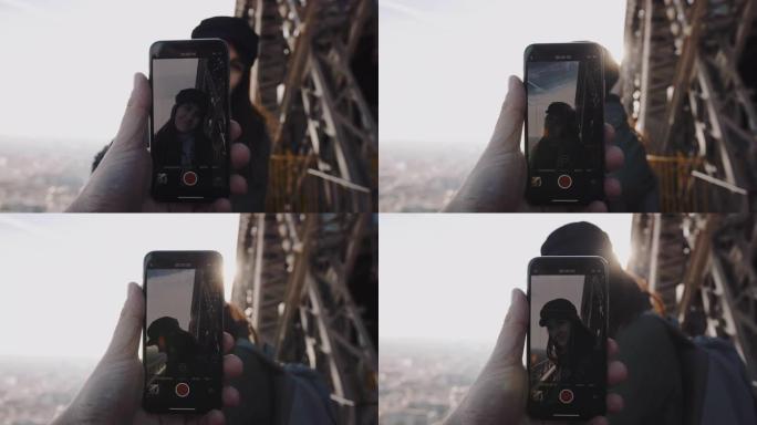 男性手中的智能手机正在拍摄艾菲尔铁塔慢动作顶部摆姿势的浪漫微笑女人的浪漫肖像。