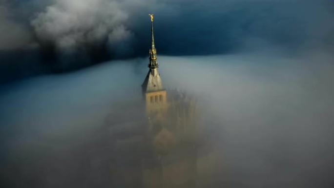 无人机在日出上飞行，圣米歇尔山城堡尖顶被浓雾覆盖，流入神秘的黑暗。