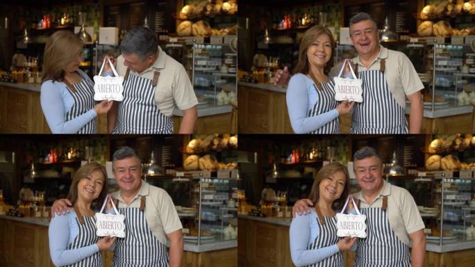 一家面包店的爱心商人夫妇老板拿着西班牙语的开放标志，对着镜头微笑