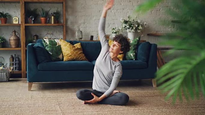 在瑜伽练习期间，漂亮的卷发女孩正伸展手臂，扭动身体坐在公寓地毯上的莲花姿势。