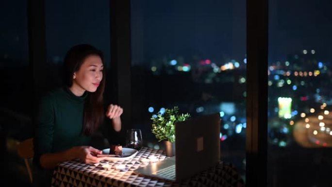 一位亚洲华裔美丽的年轻女士在魔术时刻，黄昏时在公寓里吃甜点，并使用笔记本电脑通过互联网与男友交谈