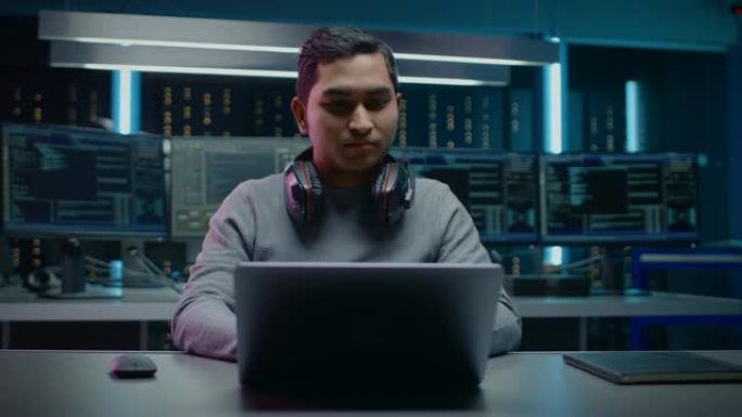 软件开发人员/黑客/程序员坐在他的办公桌前，在背景黑暗霓虹灯高科技环境中工作的笔记本电脑，有多个显示