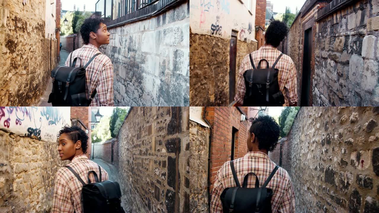 年轻的黑人妇女背着背包走在石墙之间的狭窄小巷中，向后看，跟随拍摄