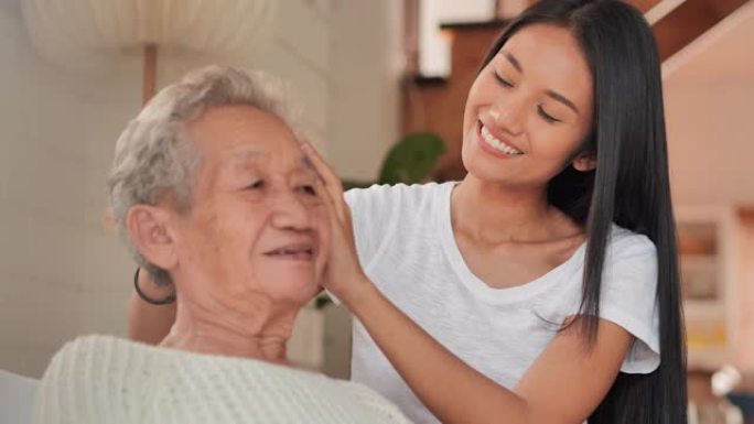 非洲女性健康访问者正在梳理她的亚洲老年女性的头发坐在沙发上，呆在家里预防冠状病毒或Covid-19.