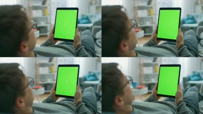 家里的年轻人躺在沙发上，使用垂直纵向模式的绿色模拟屏幕平板电脑。男人使用触摸屏设备，浏览互联网，观看