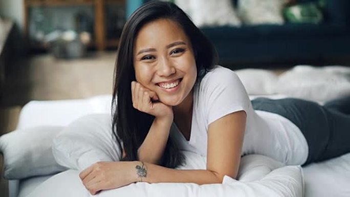 漂亮的亚洲女士躺在家里的床上，微笑着笑着看着相机的肖像。放松、好心情、快乐和情感概念。