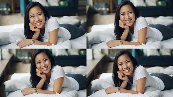 漂亮的亚洲女士躺在家里的床上，微笑着笑着看着相机的肖像。放松、好心情、快乐和情感概念。
