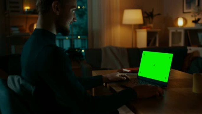 坐在办公桌前的男人在带有绿色模拟屏幕的笔记本电脑上工作。深夜在他的客厅里，男人使用笔记本电脑。侧视放