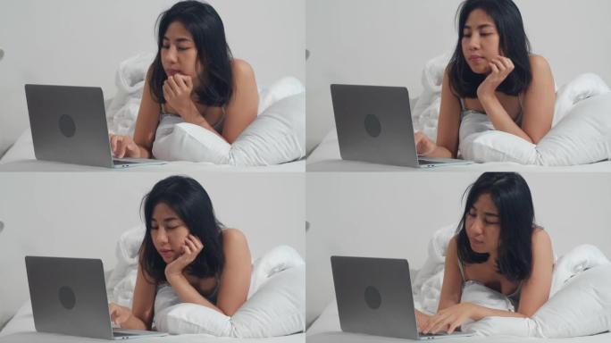 年轻的亚洲妇女使用笔记本电脑检查社交媒体感到高兴的微笑，躺在床上后早上在家里醒来，迷人的泰国女孩微笑