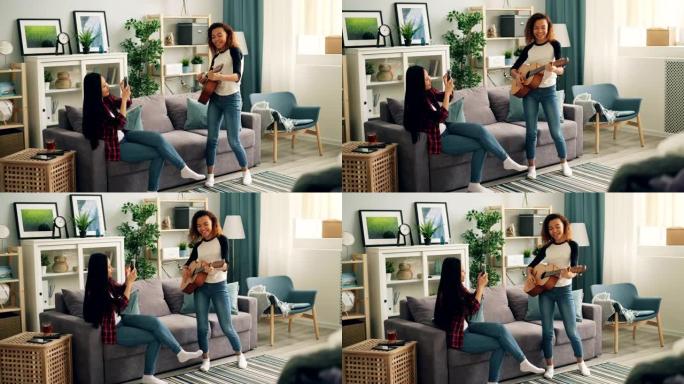 可爱的非洲裔美国妇女正在家里弹吉他，而她的亚洲朋友正在用智能手机录制视频，微笑着享受音乐和友谊。