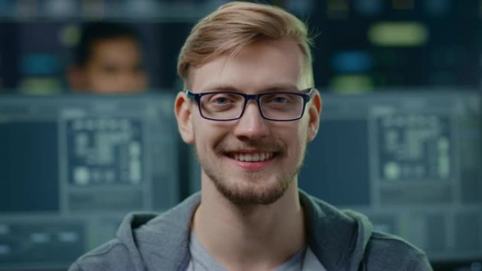 一位聪明英俊的IT专家戴着眼镜的肖像对着镜头迷人地微笑。在技术研究数据中心中带有屏幕的背景下未聚焦的
