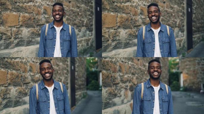 在城市的街道上，夏天，英俊的非洲裔美国男子学生微笑着看着站在户外的相机的慢动作肖像。人和幸福的概念。