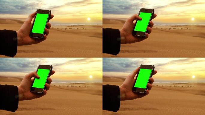 男性双手手持智能手机绿屏在日落天空的海滩上。